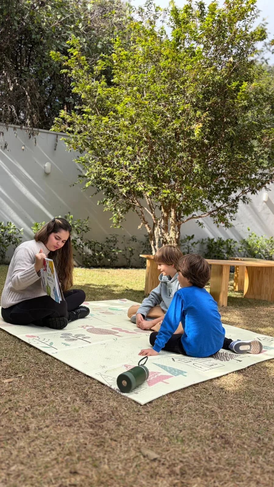 Educadora lendo um livro ilustrado para duas crianças sentadas num tapete sob a árvore